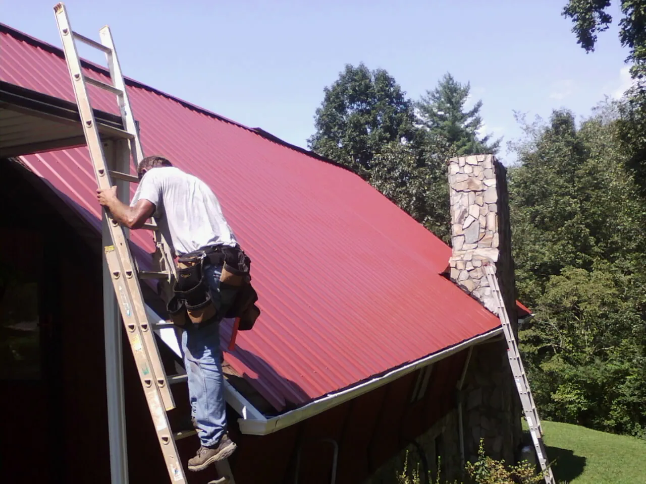 Roofer on a ladder beside a red metal roof and stone chimney — Soffit & Fascia Repair by Roofing Brothers in Mount Marion